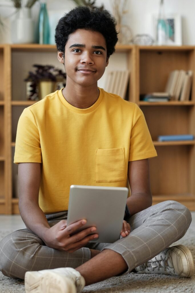 Smiling teenager holding a tablet, sitting comfortably indoors, wearing a yellow shirt.